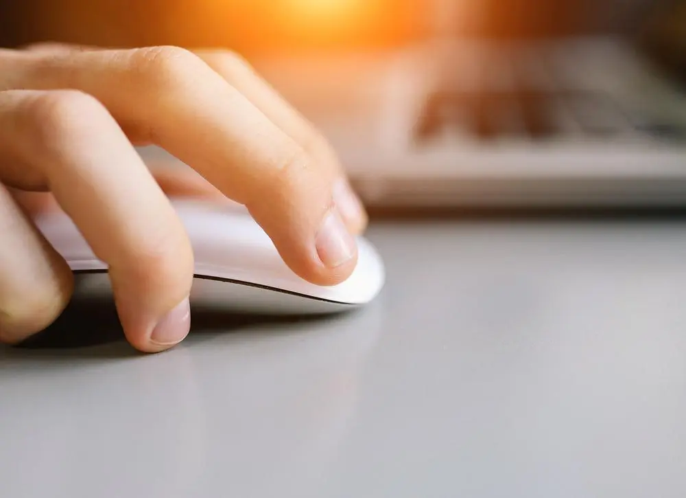 A close-up of a hand holding a white computer mouse next to a laptop on a gray surface, with warm sunlight in the background.