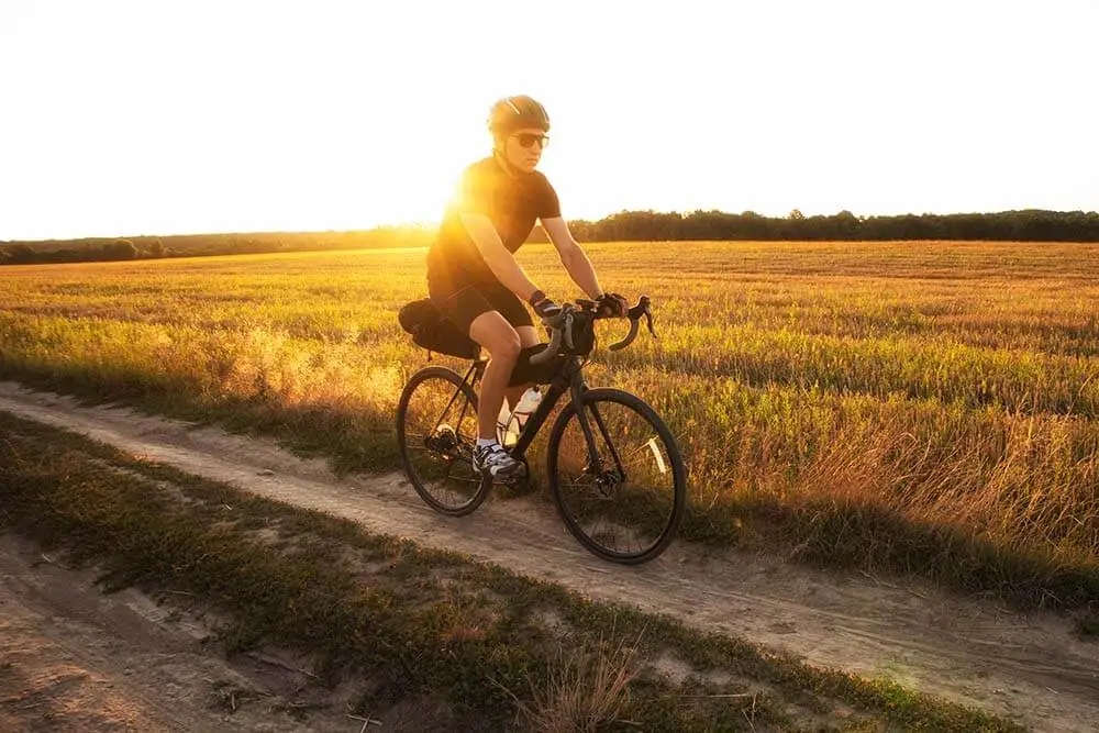 A cyclist wearing a helmet and sunglasses rides a bicycle along a dirt path through a sunlit field at sunset, with golden grass stretching into the distance.