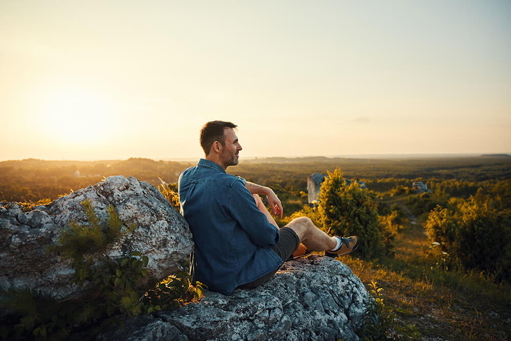 A man sits on a large rock outdoors at sunset, looking out over a scenic landscape with trees and hills, wearing a denim shirt and shorts, enjoying the peaceful view.