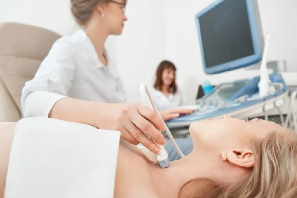 A medical professional performs an ultrasound scan on a womans neck while she lies down, with another person working at a computer in the background.