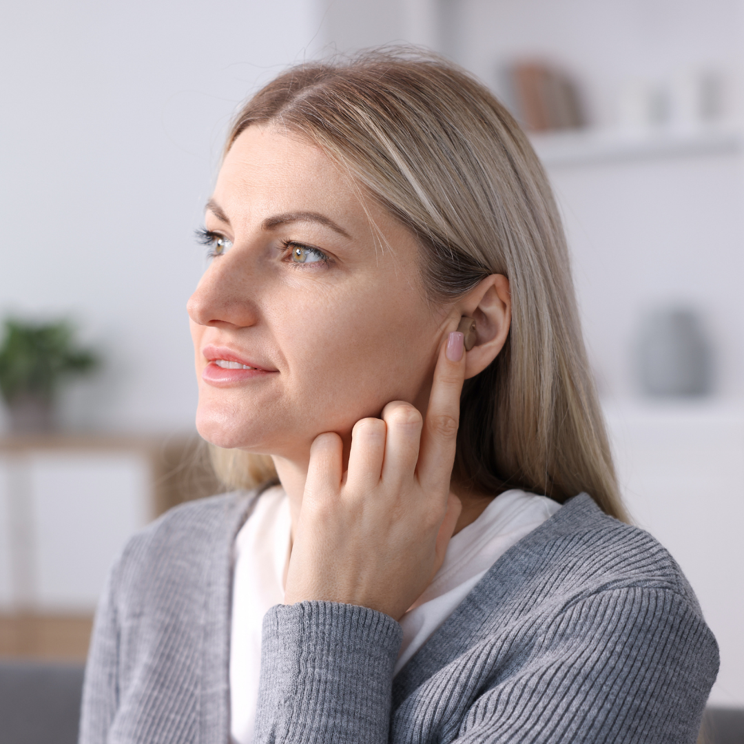 A woman with blonde hair touches her ear with her finger, appearing to focus or listen carefully. She is indoors, wearing a gray cardigan, with a blurred background of shelves and a plant.
